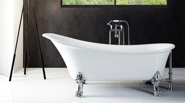 A white freestanding clawfoot bathtub with a chrome faucet, set in a modern bathroom with a dark textured wall, a black tripod floor lamp, and a two-panel window showing green trees outside.